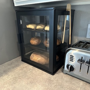 May include: A black rectangular bread box with a glass front door and three internal shelves holding various baked goods. A pair of light-colored wooden tongs hangs on the right side of the frame. The unit is positioned next to a stainless steel toaster.