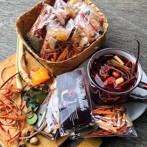 May include: A woven basket filled with clear plastic packets of dried herbs and spices, alongside a white enamel mug containing a dark red herbal infusion. A label on a packet reads Wedang Uwuh, with loose lemongrass, leaves, and ginger nearby.