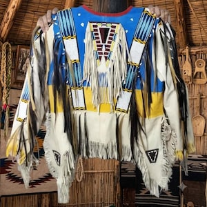 Könnte beinhalten: Eine cremefarbene Lederjacke mit langen Fransen, blauen und gelben horizontalen Bahnen sowie aufwendigen Perlenstickereien in weißen, blauen und dunkelroten geometrischen Mustern. Das Kleidungsstück hat dunkelbraune Federakzente und einen roten Kragen.