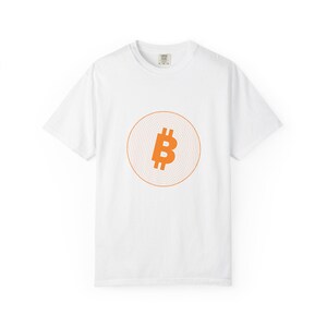 May include: A white short-sleeve t-shirt featuring a central graphic of an orange Bitcoin symbol enclosed within a circular pattern of thin, concentric orange lines. The shirt has a crew neckline and a standard fit, displaying a clean design.