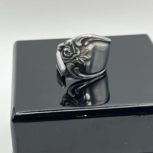 May include: A silver-toned spoon ring featuring an embossed rose motif on the front. The metal surface has a polished finish with intricate floral detailing and curved lines that wrap around the band, creating a continuous, rounded shape.