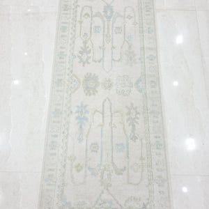 May include: A rectangular runner rug featuring a cream-colored base with faded floral and geometric patterns in soft shades of light blue and sage green. The rug has a decorative border and short, light-colored fringe along the top and bottom edges.