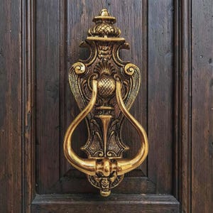 Könnte beinhalten: Ein goldfarbener Türklopfer aus Metall mit einem verzierten, symmetrischen Design, einem zentralen ananasartigen Motiv, geschwungenen Seitenarmen und einem kleinen Gesicht an der Unterseite, montiert an einer dunkelbraunen Holztür.