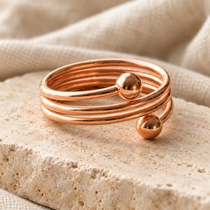 May include: A rose gold-toned metal ring featuring a multi-coil wrap design. The band consists of three continuous loops of smooth, polished wire, each ending in a small, spherical ball terminal at the open ends of the spiral structure.