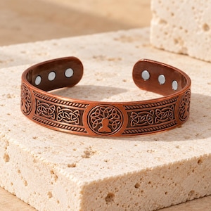 May include: A copper-colored cuff bracelet featuring an embossed tree of life motif and intricate Celtic knotwork patterns. The interior of the open-ended band displays six small, circular silver-toned magnetic inserts positioned at both ends.