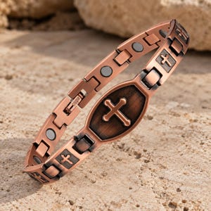 May include: A copper-toned link bracelet featuring a central shield-shaped plaque with an embossed cross. The bracelet includes alternating links with small cross motifs and circular magnetic inserts, with the word copper stamped on the clasp.
