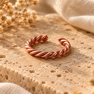 May include: A copper-colored ring featuring a twisted, rope-like design. The ring is open-ended, displaying a continuous spiral pattern throughout the band. The metallic surface has a warm, reddish-brown tone with a polished finish that reflects light.