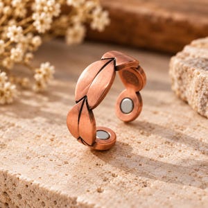 Puede incluir: Un anillo abierto de tono cobre con un diseño repetitivo en forma de hoja a lo largo de la banda. El interior del anillo muestra dos pequeños insertos magnéticos circulares de color plateado en los extremos abiertos.