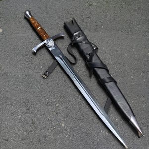 May include: A steel sword with a polished silver-tone crossguard and a rounded pommel. The handle is crafted from brown wood with a visible grain. Beside the sword lies a black leather scabbard featuring integrated black leather straps and a silver-tone tip.