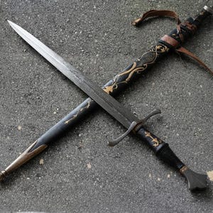Op de afbeelding: Een tweesnijdend stalen zwaard met een donker, getextureerd lemmet en een pareerstang. De zwarte schede is voorzien van goudkleurig bloemmotief, een bruine leren riem en een metalen chape. Het handvat is zwart met goudkleurige accenten.
