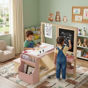 May include: A light pink and natural wood children's art station featuring a chalkboard, a paper roll holder, and integrated storage shelves. The unit includes a flat tabletop for drawing and side compartments for art supplies, designed for creative play.