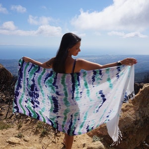 May include: A person standing on a hilltop with a view of the ocean and a distant city. They are wearing a white, blue, and green tie-dye sarong.
