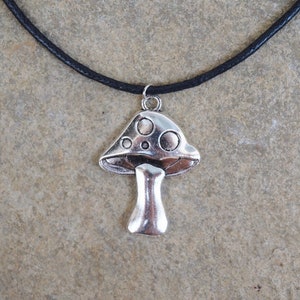 Puede incluir: Un collar con un colgante de hongo plateado en un cord&oacute;n de cuero negro. El hongo tiene un sombrero blanco con manchas negras y un tallo plateado.