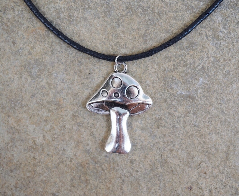 Puede incluir: Un collar con un colgante de hongo plateado en un cord&oacute;n de cuero negro. El hongo tiene un sombrero blanco con manchas negras y un tallo plateado.