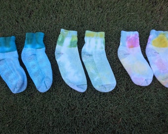 Puede incluir: Seis pares de calcetines tie-dye de colores, dispuestos en fila sobre una superficie de hierba verde. Los calcetines son todos blancos con diferentes tonos de azul, verde, amarillo y rosa.
