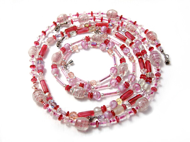 May include: A multi-strand necklace made of pink and red glass beads. The beads are of various shapes and sizes, including round, oval, and cylindrical. The necklace is strung on a silver chain.