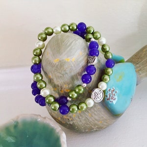 May include: Two layered bracelets with green, white, and purple beads. The bracelets feature silver Celtic knot charms.