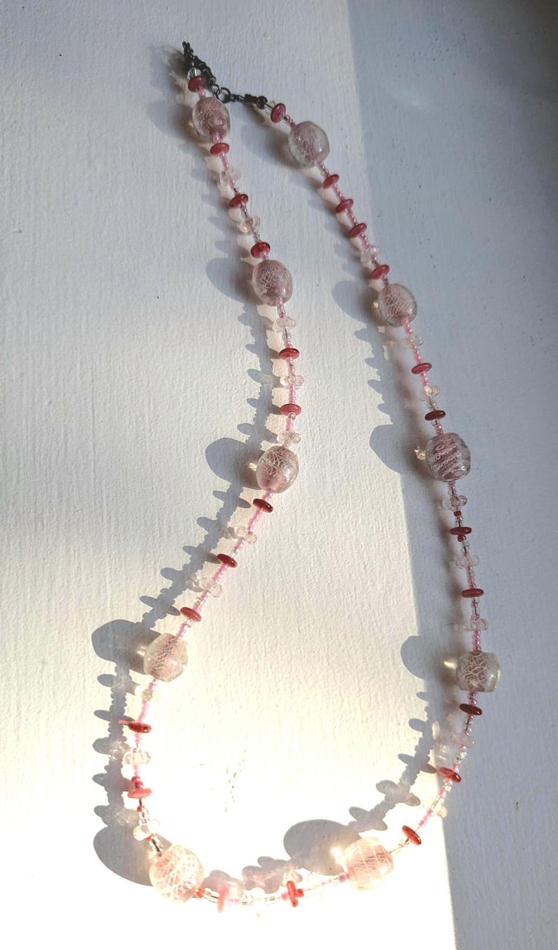 May include: A pink and clear glass beaded necklace with a silver clasp. The beads are various sizes and shapes, with some having a textured surface.