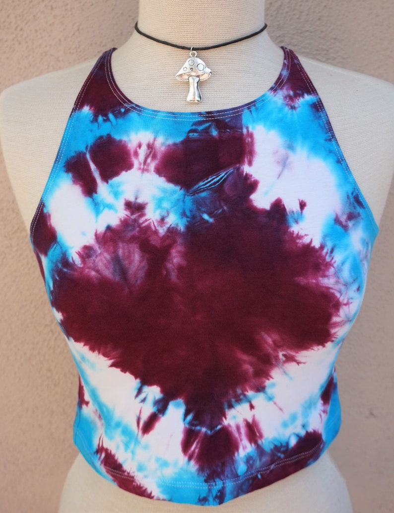 Puede incluir: Un top halter de te&ntilde;ido anudado en tonos de blanco, azul y burdeos. El top es corto y tiene un ajuste holgado. Un colgante de plata en forma de hongo cuelga de un collar gargantilla negro.