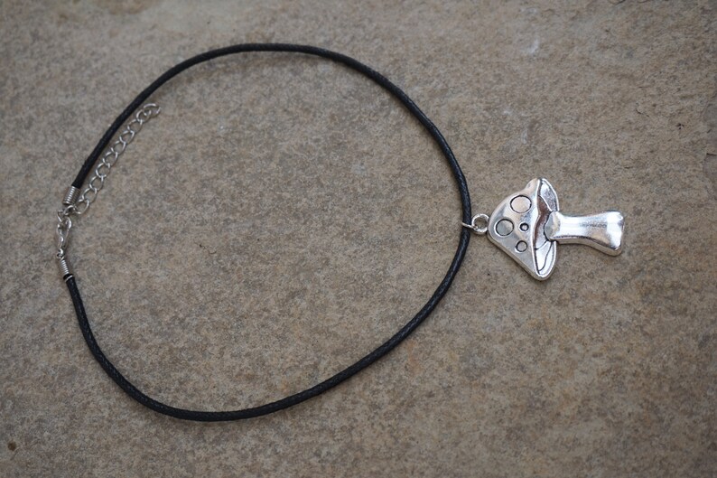 Puede incluir: Un collar de cord&oacute;n de cuero negro con un colgante de hongo plateado.