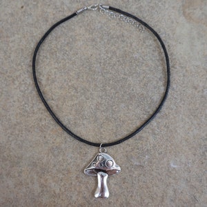 Puede incluir: Un collar de cord&oacute;n de cuero negro con un colgante de hongo plateado. El hongo tiene una parte superior abovedada con una superficie texturizada y un tallo.