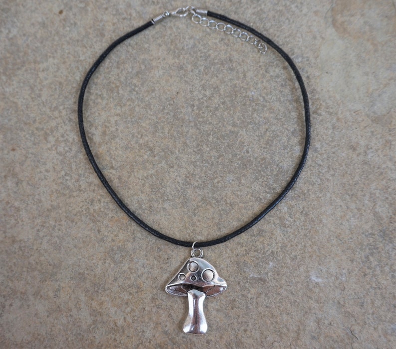 Puede incluir: Un collar de cord&oacute;n de cuero negro con un colgante de hongo plateado. El hongo tiene una parte superior abovedada con una superficie texturizada y un tallo.
