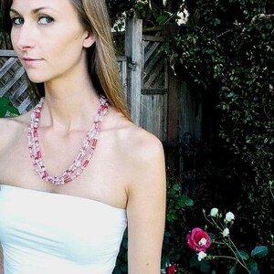 May include: A woman wearing a white strapless top with a layered pink and white beaded necklace. The necklace is made of small, round beads and is worn in a multi-strand style.