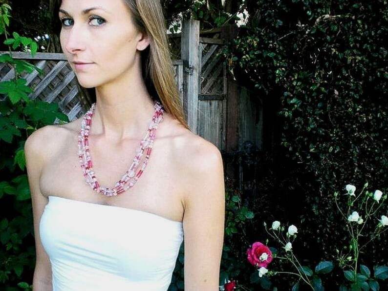 May include: A woman wearing a white strapless top with a layered pink and white beaded necklace. The necklace is made of small, round beads and is worn in a multi-strand style.