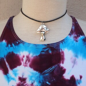 Puede incluir: Un collar con un colgante de hongo plateado en una gargantilla negra. El collar se lleva sobre un top de tirantes te&ntilde;ido anudado con colores azul, morado y blanco.