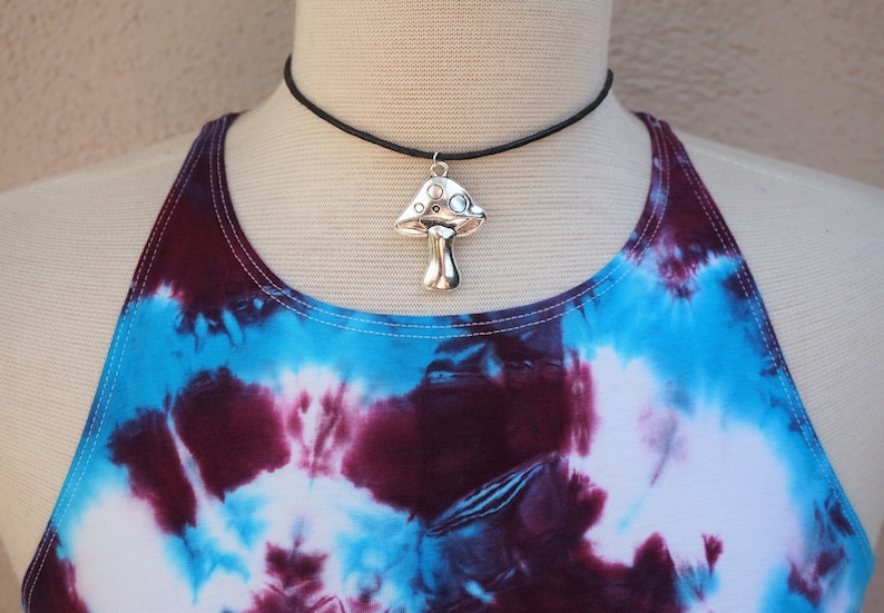 Puede incluir: Un collar con un colgante de hongo plateado en una gargantilla negra. El collar se lleva sobre un top de tirantes te&ntilde;ido anudado con colores azul, morado y blanco.
