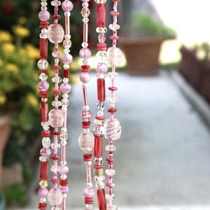 May include: A multi-strand necklace made of pink, red, and clear glass beads. The necklace features a variety of bead shapes and sizes, including small round beads, larger round beads, and long, thin beads.