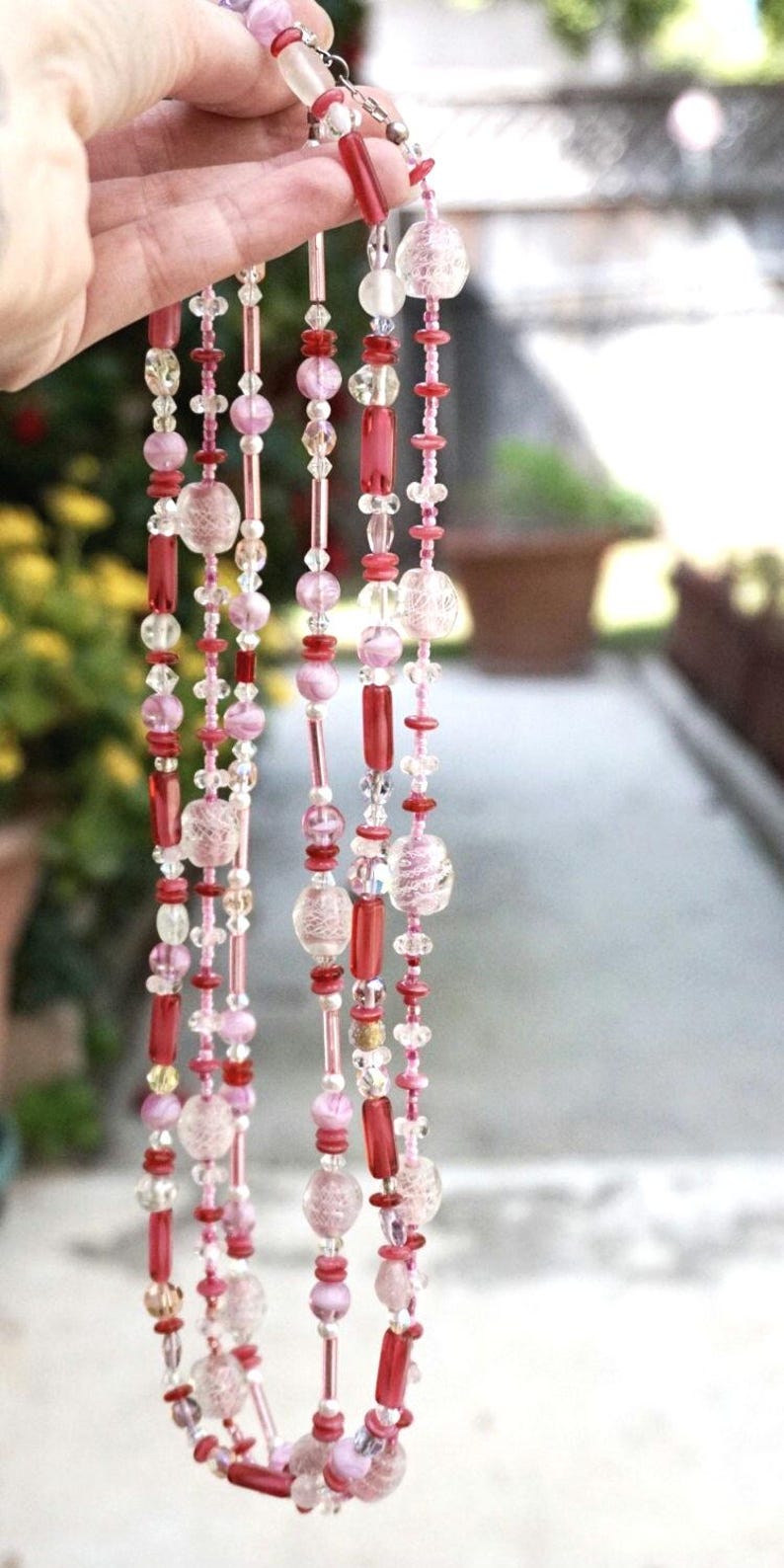 May include: A multi-strand necklace made of pink, red, and clear glass beads. The necklace features a variety of bead shapes and sizes, including small round beads, larger round beads, and long, thin beads.