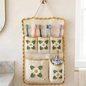 May include: A cream-colored crochet wall organizer with a mustard yellow scalloped border. It features five pockets, each decorated with a white and yellow daisy appliqué and green leaves. The organizer holds six toothbrushes and a soap dispenser.