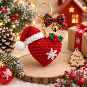 May include: A red crocheted heart keychain featuring a white-trimmed red Santa hat, a white snowflake embellishment, and a green holly leaf accent. The keychain includes a gold-tone metal ring, a plaid fabric bow, and a small gold jingle bell.