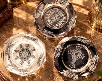 May include: Three faceted glass ashtrays with octagonal rims. Two are clear with black printed cross and floral motifs, while one is dark blue with a white printed cross design. Each features intricate circular patterns and ornate detailing.