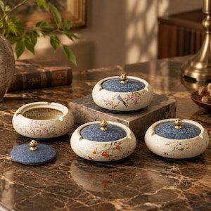 May include: A collection of round ceramic ashtrays with cream-colored bases featuring floral and botanical illustrations. Each ashtray includes a textured blue lid topped with a small gold-toned metal knob. Some lids are removed to show the interior.
