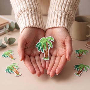 Peut inclure: Un écusson brodé en forme de palmier avec un tronc marron et des palmes vert vif bordées de blanc. Le motif présente des détails de broderie texturés, illustrant une plante tropicale qui est répétée sur plusieurs écussons identiques.