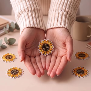 Könnte beinhalten: Eine Sammlung von Sonnenblumen-Aufklebern mit einer dunkelbraunen Mitte in Gitteroptik, umgeben von leuchtend gelben und orangefarbenen Blütenblättern. Die Aufkleber haben einen weißen Rand und sind in einer Gruppe angeordnet.