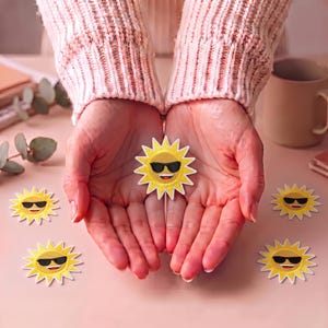 Puede incluir: Una colección de cinco parches bordados de sol, cada uno con un sol amarillo brillante con una cara sonriente y gafas de sol negras. Los parches tienen un borde blanco y están dispuestos con uno centrado y cuatro alrededor en una superficie.