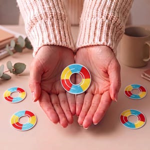 Puede incluir: Un parche circular con un diseño segmentado que presenta secciones alternas de hilo rojo brillante, amarillo y azul claro. El bordado está dispuesto en un patrón de anillo con un borde blanco, y varios parches idénticos están esparcidos.