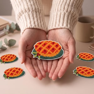 May include: An embroidered patch featuring a lattice-topped fruit pie on a dark green plate. The pie has a golden-brown crust with a red fruit filling visible through the lattice pattern. Small green leaves and red berries decorate the plate edge.