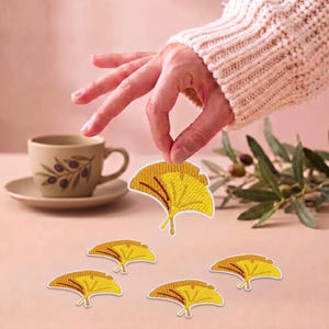 Op de afbeelding: Een set van vijf geborduurde ginkgo-blad patches in levendig geel met bruine aders. Elke patch heeft een waaiervormig ontwerp met een opvallende ingekeepte rand en is afgewerkt met een strakke, witte randafwerking rondom het borduurwerk.