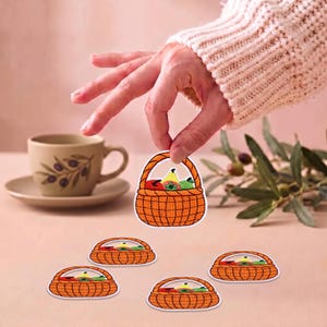 Puede incluir: Un conjunto de parches bordados, cada uno con forma de cesta naranja tejida llena de frutas coloridas, incluyendo piezas rojas, amarillas y verdes. Los parches presentan un patrón de cuadrícula en la cesta y un asa curva en la parte superior.