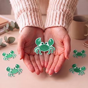 May include: A collection of small, embroidered crab patches featuring a vibrant green color with a white border. Each patch depicts a stylized crab with two raised claws, six legs, and two small black eyes, designed for application onto fabric surfaces.