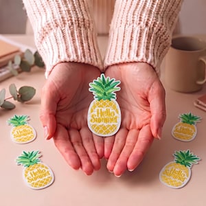 Puede incluir: Un parche bordado de piña en colores amarillo y verde con el texto Hello Summer en letra cursiva. El parche tiene un borde blanco y está rodeado por otros cuatro parches de piña idénticos sobre una superficie de tono claro.