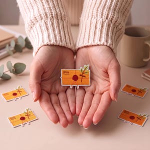 Pode incluir: Um emblema bordado em forma de envelope retangular laranja com um selo de cera vermelho, um pequeno ramo verde e linhas horizontais que representam um selo postal. O emblema tem uma borda branca e é exibido num fundo neutro.