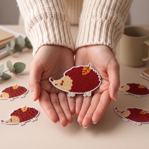 May include: An embroidered patch shaped like a hedgehog with a dark red body, a tan face, and a small yellow leaf on its back. The patch features a white border and detailed stitching, showcasing a stylised animal design with a pointed snout.