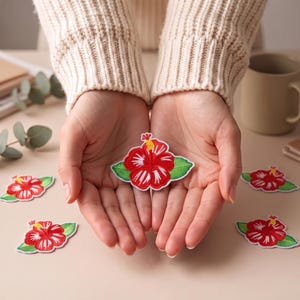 May include: An embroidered hibiscus flower patch featuring vibrant red petals with white highlights, a yellow stamen, and green leaves. The patch is shaped like a blooming flower and is designed for application onto fabric surfaces as a decorative accent.