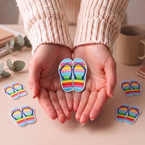 Può includere: Una coppia di toppe ricamate a forma di infradito con strisce orizzontali arcobaleno nei colori rosso, arancione, giallo, verde, blu e viola, dotate di cinturini blu scuro. Le toppe presentano un bordo bianco e sono mostrate insieme a vari pezzi identici.