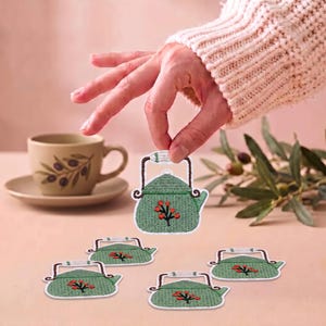 May include: A set of embroidered patches featuring a green teapot design with a brown handle and a small red berry branch motif. The patches have a white border and are shaped like a classic stovetop kettle with a spout and a top handle.